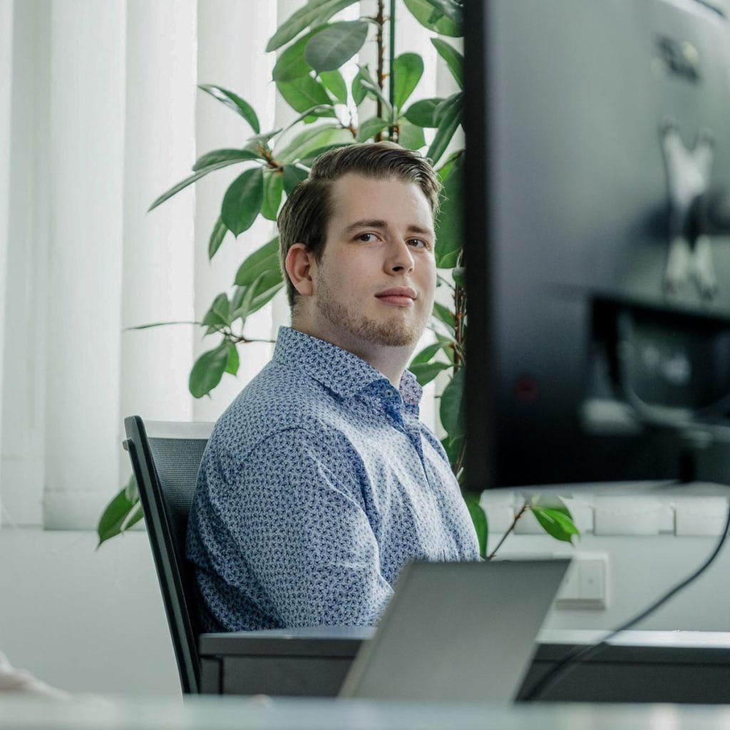 Ein junger Mann im Hemd sitzt an seinem Arbeitsplatz und blickt direkt in die Kamera. Das Bild ist Teil der Content-Produktion, die auf Authentizität setzt, um die Arbeitsatmosphäre und das Team von Dinape authentisch und professionell darzustellen.